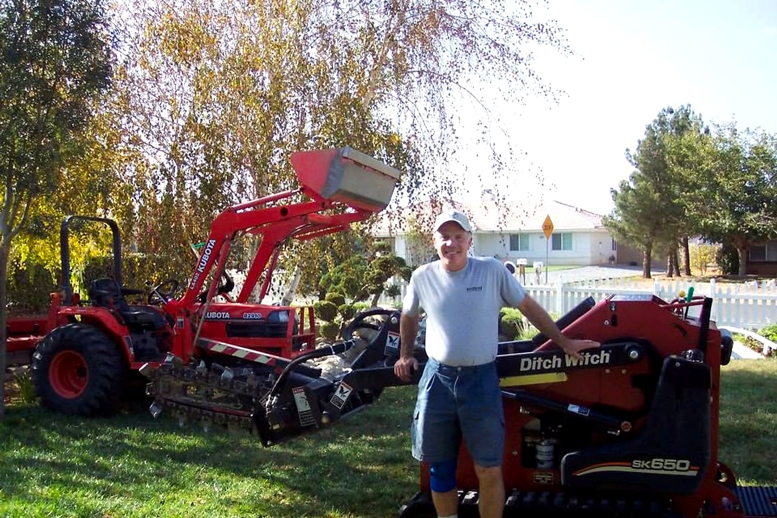 Gary Swank standing between his red Kubota tractor and his Ditch Witch mini-skid with a trencher